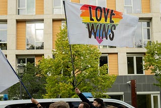 A city street with someone holding up a white flag with a rainbow heart on it, reading “LOVE WINS.”