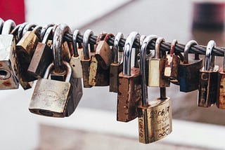 This image shows a collection of various old padlocks attached to a metal wire, illustrating a display of love locks. The padlocks are in different shapes, sizes, and states of wear, some rusty and weathered, others still shiny. Several locks have inscriptions, suggesting that they were placed there by individuals or couples to symbolize their love and commitment. The background is blurred, focusing attention on the details and textures of the locks.