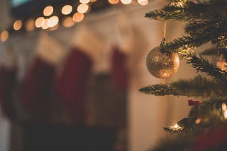 A gold, sparkly ornament hanging from a Christmas tree. Stockings and lights blurred in the background, over a fireplace.