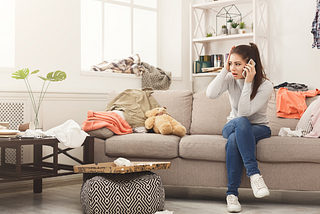 Woman, sitting on a cluttered sofa, with her phone to one ear and a shocked expression on her face.