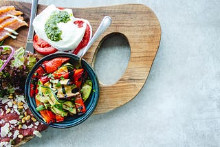 a cutting board with healthy foods such as avocado, tomato and mozzarella sandwich, greens.