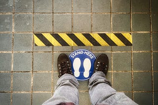 View down on feet standing on a “stand here” mark, behind a black and yellow warning line.