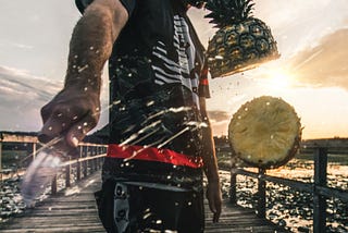 Man on dock at sunset slices pineapple with knife