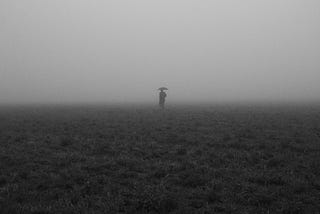 A monochromatic picture of a man in dark clothes, surrounded by heavy fog, holding an umbrella over his head whilst walking in a field of grass.