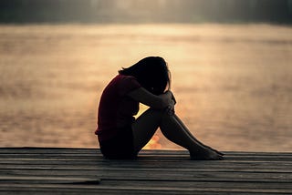 A young woman sitting on a wooden deck with her head and hands on her knees.