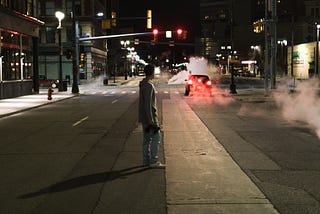 Young man standing in deserted city street, watching glowing red tail lights flashing in the distance.