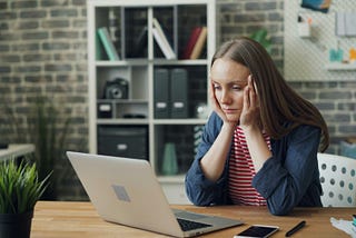 Photo of a sad looking woman with her head in her hands working at a laptop