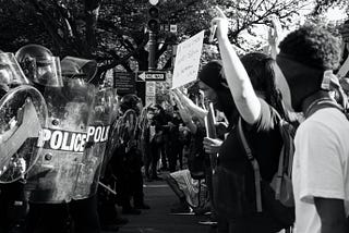 A black and white photo of police with shields facing protesters.