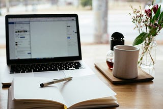 A journal and pen sitting on a desk in from of a laptop screen and a mug of coffee.