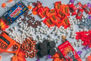 Spread out on a table there’s halloween candy and decorations with marshmellows, pretzels, oreos and raisonets sprinkled on the table too