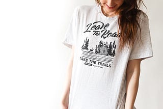 A woman wearing a white t-shirt that says “Leave the Road Take The Trails”