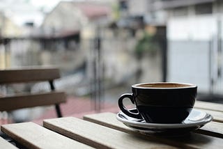 A coffee cup and saucer on a wooden table. A wooden can be seen in the background, and there are some buildings in the background, but the background is blurred.