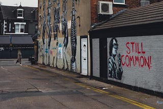A desolate side-street with graffiti on the walls. Beside the image of Pulp singer Jarvis Cocker there is a caption which reads ,’Still Common’.