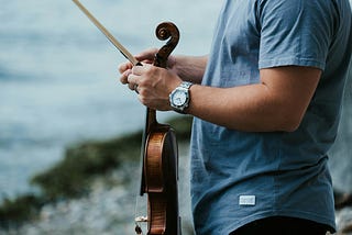 Wearing a watch on left wrist, a man in a blue shirt holds a violin and a bow.