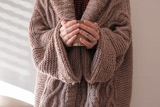 Wrapped in a cozy, thick brown knit sweater, a woman holds a steaming mug in her hands.
