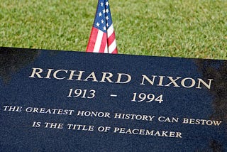 A black marble plaque with Richard Nixon’s name with the timespan 1913–1994, with the words “THE GREATEST HONOR HISTORY CAN BESTOW IS THE TITLE OF PEACEMAKER” below it. Green grass and American flag in the background.