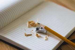 pencil sharpener and pencil lying on an open lined notebook. The pencil looks like it’s just been sharpened; there are shavings next to it.