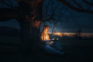 A young man sitting by a tree at night and reading a book
