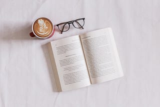 Fotografia de um livro aberto, na parte de cima um oculos com armação preta e no lado esquerdo uma xicara de café.