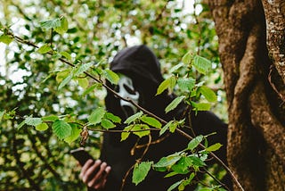 Guy in a scream mask, standing behind a tree and checking his phone