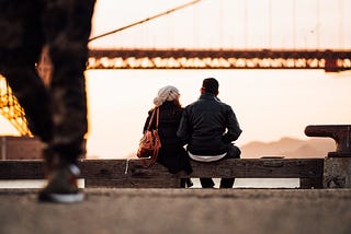 A guy and a girl sitting closely on the edge of a road in front of river.