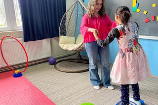 A 10-year-girl balances on a therapeutic board holding hands with a young woman standing across from her. There is a blackboard with magnets of different sizes and geometric shapes to their right. A self-standing hammock in the shape of an egg stands in the corner for swinging.