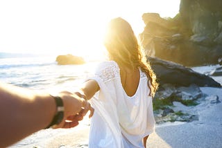 a couple on the beach, man has his arm stretched out as the woman holds on to his hand, her back turned watching the waves.