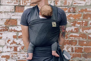 A father is holding his son in a baby sling attached to the front of his body. The father has colourful tattoos on his right arm. Baby has dark hair and bare feet.