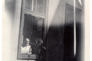 Black and white photograph in portrait orientation of a wall of a building with two windows next to each other. Both windows are divided into three parts by window frames; two small windows at the top and one larger window at the bottom. The bottom part of the window on the left is open. A man in a white shirt leans on the window frame in the bottom left corner, only his top half is visible. To his right is a potted plant. At the top of the image is a handwritten annotation in blue ink.