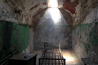 A old prison cell with a bare bed and light entering from above through a tiny window.