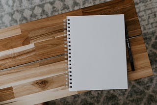 A wooden tabletop with a spiral bound notebook. Next to the notebook is a pen.