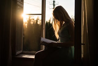 The silhouette of a woman sitting on a windowsill reading a book. Golden sunlight is coming in through the open window.