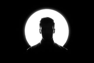 Black and white photo. A man, his face in shadow, stands in front of a giant moon.