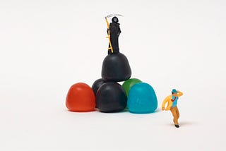 A tiny black-robed figure of Death holding a scythe stands on top of what looks like black, red, green, and blue sugarplums. A tiny figure in the foreground looks like a man in a coat, tie, and hat running from Death.