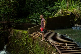 A white bearded man in red shorts and a tank top stands on top of a rock dam covered with moss. There is a wide wooden ladder lying sideways on top of the dam.