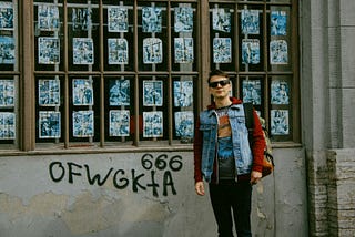 Young man wearing sunglasses, a red hoodie with design partially hidden by a denim vest, black jeans, and a backpack. He is standing next to a wall with a large window full of promotional posters. Beneath the wall is spray-painted 666 and random letters.