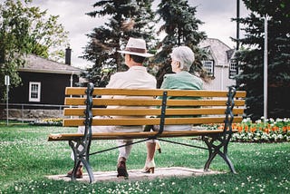 older man and woman sitting on a park bench with their back to the camera