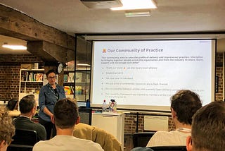 Me standing at a podium in front of a big screen at the Made Tech offices, pulling a funny face and introducing our Community of Practice