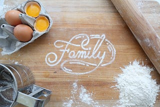 family written in flour on wooden board with eggs, flour, sifter, and rolling pin