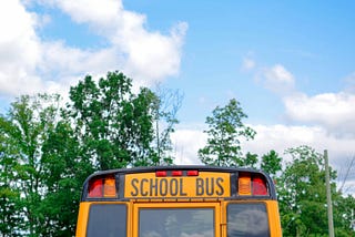 This image shows the rear top section of a traditional yellow American school bus, prominently displaying the words “SCHOOL BUS” in black on a background field of yellow. Above the text are two red lights. The sky is partly cloudy and trees can be seen in the background, indicating a daytime setting.