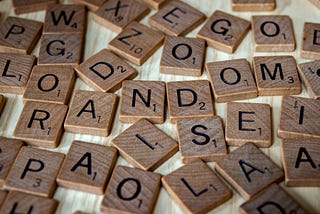 An array of Scrabble pieces (single letters with subscript point values) sit scattered across a wooden table, mid-game.
