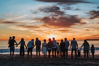 people standing on beach in a line, backs to the camera gazing at the sun set