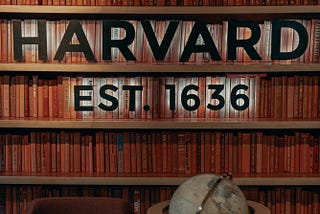 A bookcase with the words “Harvard” and “Established 1636” in front of two rows of books, with an armchair and a globe in front of the bookcase.