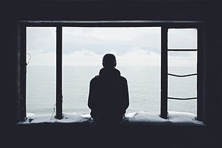 A person sits on the edge of a stone window opening, flanked by vertical metal bars on either side. They look out onto an ocean view. Clouds are gathering in the distance.