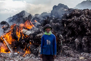 Man standing in front of a burning heap of rubbish, in a large open landfield