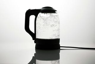 A transparent kettle with black plastic handle, lid and base, is filled with boiling water.