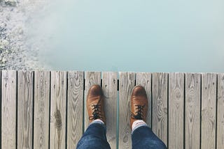 Person looking down at their shoes as they stand on wooden walkway over cloudy water.