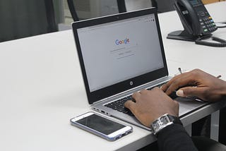 Google, shown on the screen of a laptop. A man wearing a watch. There is an iPhone on the table.