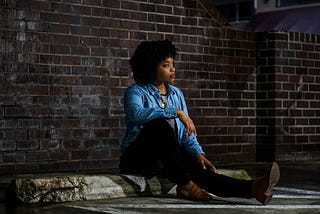 A woman with dark, curly hair wearing a denim jacket, dark pants and low-heeled shoes sits by the curbside with her right leg bent and right elbow rested on her knee. With her head turned to her left, she seems lost in thought.