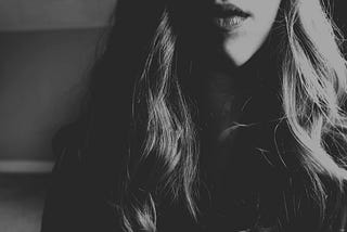 Black and white photo of the bottom half of a woman’s face from the top of her lips to just below her shoulders. Her blonde wavy hair falls over her shoulders and she’s wearing lipstick. She’s not smiling.